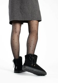 Botines de ante negro con forro de pelo sintético, punta redondeada y cordones negros. La persona lleva medias negras con lunares y un vestido gris.