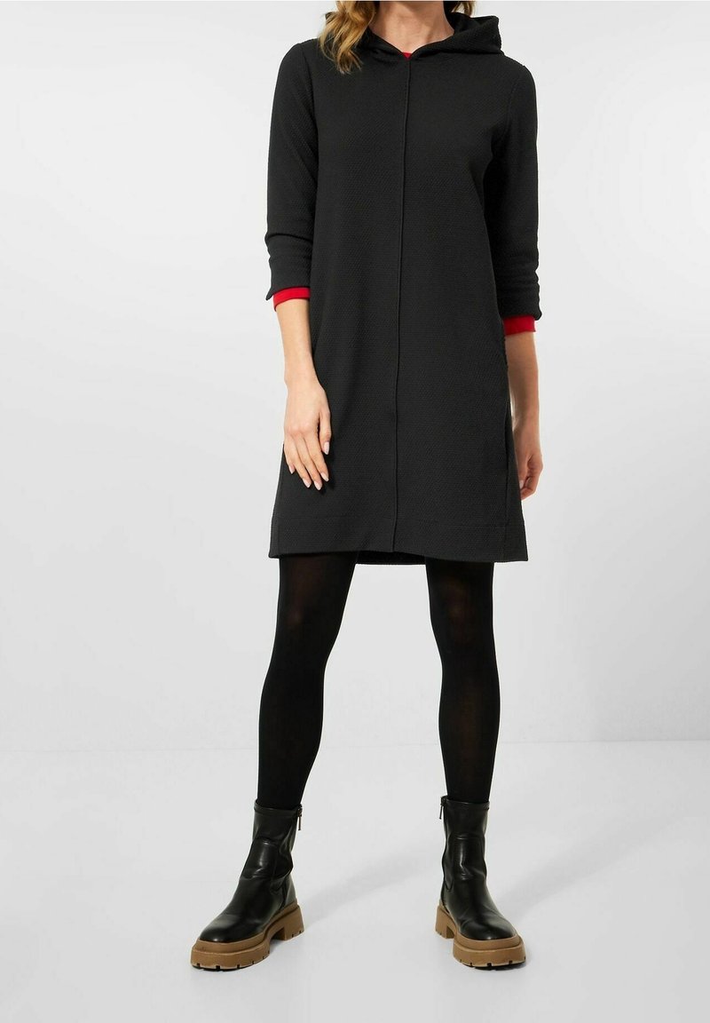 Robe noire à capuche avec manches trois-quarts, poignets rouges et tissu texturé. Associée à des bottines noires et des collants foncés.