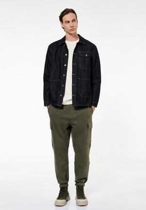 Hombre de pie con las manos en los bolsillos, lleva una chaqueta vaquera negra, camisa blanca, pantalones cargo oliva y botas de senderismo beige sobre fondo blanco.