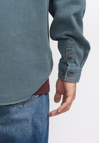 Lange mouwen knoopshirt in teal stof met een gestructureerde weving, met bruine knopen bij de manchet, gedragen over een bordeauxrood shirt en spijkerbroek.