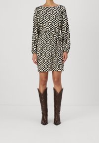 Eine Frau trägt ein beiges und schwarzes, geometrisch gemustertes Kleid mit einem Gürtel und braunen knielangen Lederstiefeln vor einem schlichten Hintergrund.