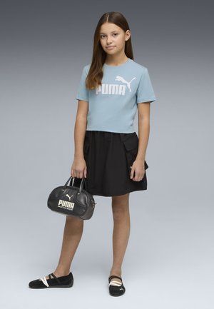 Niña con camiseta azul claro de Puma, falda negra y zapatos negros de Puma, sosteniendo un bolso negro de Puma, de pie contra un fondo gris.