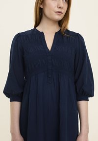Dunkelblaues Kleid mit gefälteltem Oberteil, V-Ausschnitt und puffärmel. Weicher, strukturiertes Stoff mit fließender Silhouette.