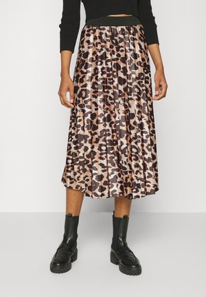 Person, die einen plissierten Midi-Rock mit Leopardenmuster, einen schwarzen, cropped Pullover und schwarze, klobige Plateau-Stiefel trägt, steht vor einem schlichten Hintergrund.