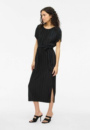 Robe midi plissée noire à manches courtes, taille ceinturée et fentes latérales. Texture lisse avec de légères lignes verticales. Sandales noires à talons.