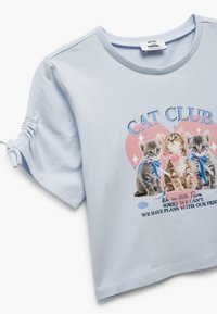 Camiseta de algodón azul claro con mangas cortas y detalle fruncido. Presenta un gráfico de tres gatos con el texto: "CLUB DE GATOS" y frases divertidas.