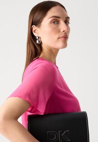 Svart läderväska med "DK" präglat, hålls av en person i en rosa genomskinlig blus. Örhängen är runda och metalliska, vilket kontrasterar med outfiten.