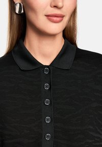 Schwarzes Poloshirt mit strukturiertem Design, ausgestattet mit einem Kragen, sechs schwarzen Knöpfen und dezenten Mustern. Das Model trägt silberne Ohrringe.
