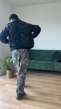 Jaqueta bombardeiro preta com detalhes franzidos nas costas, combinada com calças de camuflagem cinzas. Ambiente interno com sofá verde e planta em vaso.