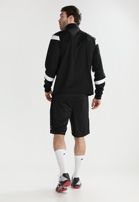 Schwarze Sportjacke mit weißen Schulterstreifen, kombiniert mit schwarzen Shorts. Modell trägt weiße Socken und schwarz-rote Sportschuhe und steht mit dem Rücken zur Kamera.