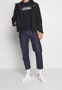 Svart huva jacka över en marinblå t-shirt med tryck, kombinerat med korta blåjeans och vita sneakers. Vita strumpor synliga ovanför skorna.