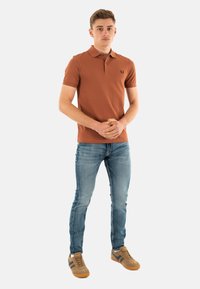 Bruine polo met korte mouwen, logo op de linkerborst, gecombineerd met lichtblauwe skinny jeans en bruine sneakers met donkere accenten.