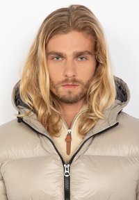Lichtbeige pufferjack met capuchon, voorzien van een zwarte rits, textuurstof, en een gebreide crèmekleurige kraag eronder.
