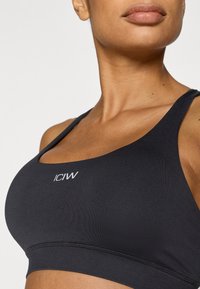 Μαύρο αθλητικό σουτιέν με στρογγυλή λαιμόκοψη, κατασκευασμένο από λείο ύφασμα. Διαθέτει το λογότυπο "ICIW" σε λευκό και υφασμένη κάτω ζών η. Ρυθμιζόμενοι ιμάντες.