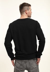 Uomo con capelli corti e orecchini, che indossa una maglietta nera a maniche lunghe e jeans grigi strappati, che volta le spalle su uno sfondo bianco.