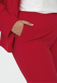 Röda skräddarsydda byxor med en slät textur, utrustade med sidofickor och en figursydd design, kombinerade med en vit topp och röd jacka.