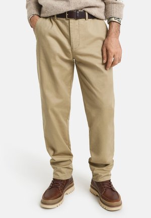 Beige cotton trousers with a relaxed fit, front pleat, and a dark brown belt. Paired with brown boots featuring a textured sole.
