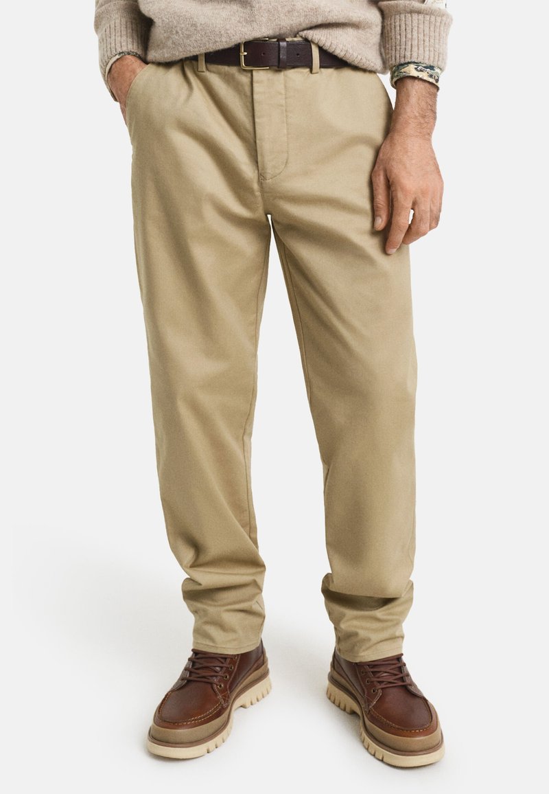 Pantalon en coton beige avec une coupe décontractée, pli avant et une ceinture marron foncé. Associé à des bottes marron avec une semelle texturée.
