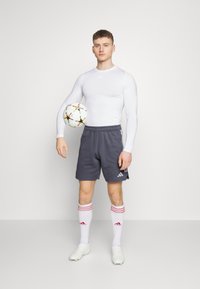 Sportkleding met een nauwsluitend wit longsleeve shirt, grijze shorts, witte sokken met rode strepen en witte voetbalschoenen, terwijl hij een voetbal vasthoudt.