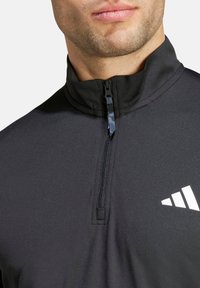 Parte superior negra atlética con cremallera, con un alto cuello, hecha de un material suave. Presenta un pequeño logo blanco y un cierre de cuello con cremallera.