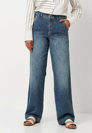 Wide-leg blue denim jeans with a slightly faded finish, featuring a front button closure and pockets, paired with a striped shirt and sandals.