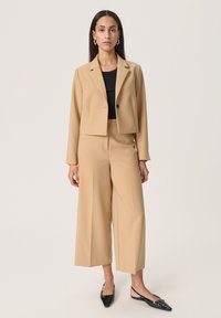 Blazer beige accorciato con un singolo bottone nero e pantaloni ampi abbinati, abbinati a décolleté nere a punta, su uno sfondo neutro.
