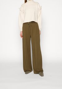 Ljusbeige korttröja med puffärmar och dekorativa knappar, tillsammans med olivgröna vida byxor och grå slip-on skor.