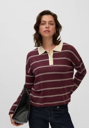 Jonge vrouw met kort bruin haar draagt een bordeaux gestreept shirt met lange mouwen en donkere jeans, en houdt een zwarte handtas aan haar linkerarm.