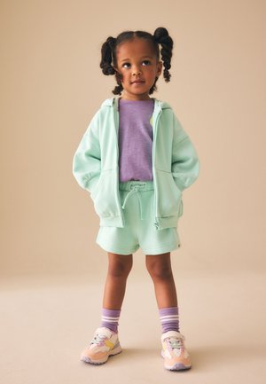 Jauna mergina stovi su kišenėmis mintinės žalsvos spalvos džemperyje su gobtuvu, derančiais šortais, violetine palaidine, dryžuotomis violetinėmis kojinėmis ir spalvingais sportbačiais.