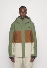 Chaqueta color bloqueada en verde, marrón y beige. Cuenta con capucha, cierre frontal con cremallera y puños elásticos. Está hecha de un material duradero y resistente a las inclemencias del tiempo.
