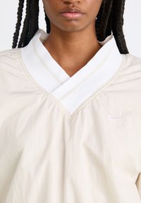 Ανοιχτόχρωμο μπλουζάκι σε κρέμα από ελαφρύ υλικό, με διπλή λευκή κολάρο που έχει δύο λεπτές μπεζ ρίγες. Το λογότυπο της Nike κεντημένο στο στήθος.