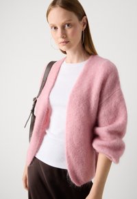 Cardigan rosa soffice realizzato in materiale morbido, con una vestibilità ampia, maniche larghe e design aperto, abbinato a una maglietta bianca e pantaloni marroni.