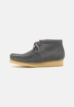Stivale alla caviglia in suede grigio con punta rotonda, chiusura con lacci e suola in gomma naturale. Presenta dettagli cuciti lungo i bordi.