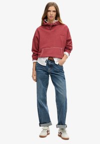Sudadera roja con capucha y bolsillo frontal, llevada sobre una camiseta blanca, combinada con jeans azules y zapatillas blancas. Estilo casual, corte relajado.