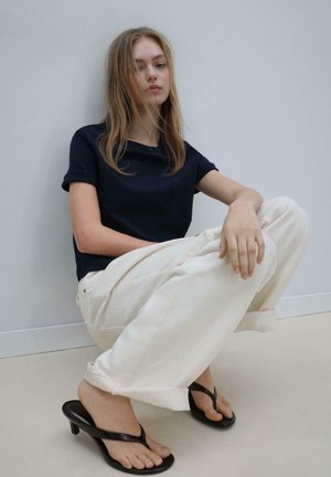 Young woman with long hair squatting, wearing a black short-sleeve top, loose white pants, and black heeled flip-flops against a plain wall.