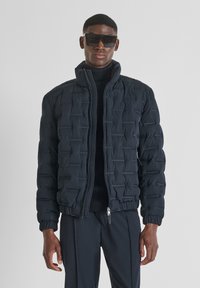 Uomo in giacca trapuntata nera, dolcevita nero, pantaloni neri e grandi occhiali da sole rettangolari neri che sta in piedi contro uno sfondo semplice.