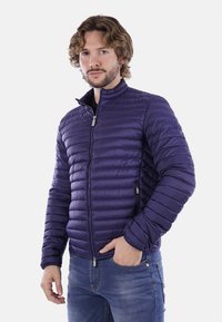 Giacca imbottita di colore viola scuro, con colletto alto, maniche lunghe e zip frontale, abbinata a jeans in denim. Superficie testurizzata con cuciture orizzontali.