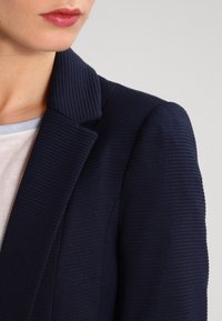 Marinblå strukturerad kavaj med slagkrage och ribbat tyg. Nära bilden visar detaljerna på kragen och axelsömmen.