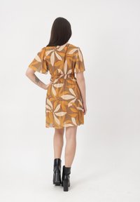 Mujer con cabello largo y oscuro, vestida con un vestido de manga corta y cintura ajustada con patrones de hojas en color naranja y beige, botas negras de tacón hasta el tobillo, de pie con la espalda visible.