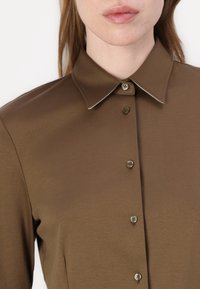 Brown collared shirt with a smooth texture, featuring a button-down front and contrasting white stitching along the collar.