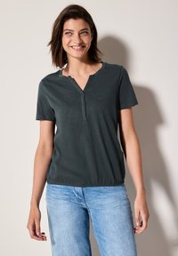 Chemise à manches courtes vert foncé avec un col en V et un ourlet élastique, fabriquée en tissu doux, associée à un jean bleu clair.