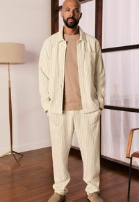 Striped, cream-colored jacket with a button front and patch pocket, paired with matching tapered pants and a beige t-shirt. Soft texture.