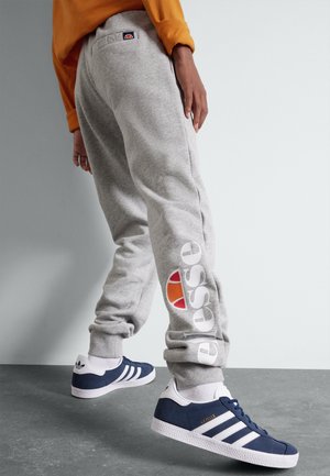 Person iført grå Ellesse joggingbukser og marineblå Adidas sneakers, stående på en grå gulv mod en hvid væg.
