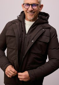Dunkelbraune Steppjacke mit Reißverschluss, zwei Brusttaschen und Kapuze. Über einem strukturierten cremefarbenen Pullover getragen.