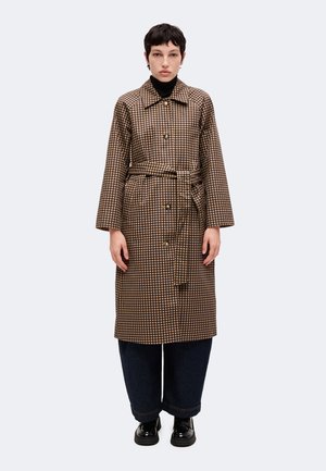 Personne portant un long manteau marron à carreaux avec une ceinture, un col roulé noir, un pantalon large foncé et des chaussures noires, debout devant un fond uni.