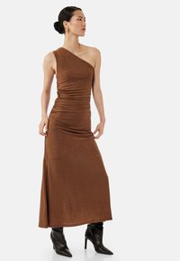 Brązowa sukienka na jedno ramię z połyskliwej, elastycznej tkaniny. Posiada marszczone detale i dopasowany krój, zestawiona z czarnymi kozakami do kolan.