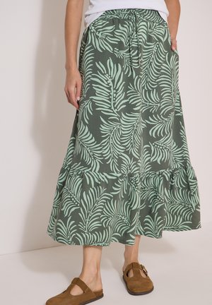Mujer con una falda midi verde con estampados de hojas verde claro, cintura elástica y zapatos marrones de ante sin cordones con detalle de hebilla.