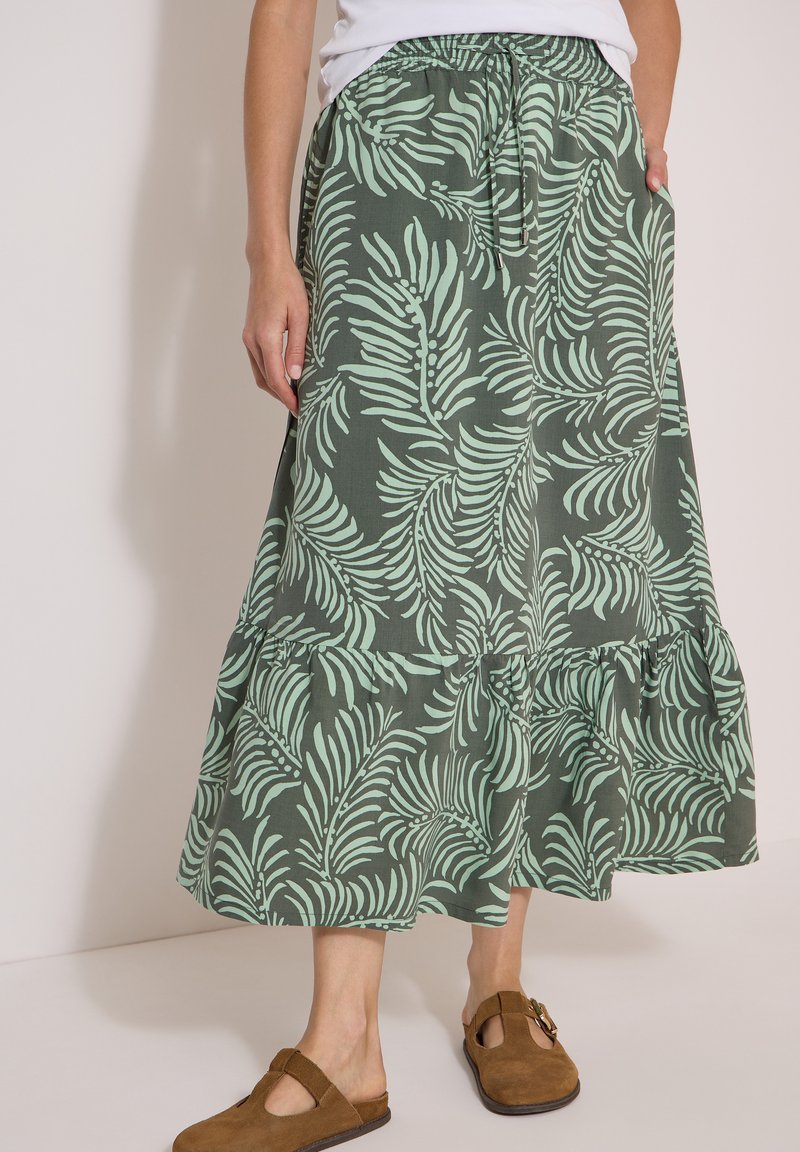 Femme portant une jupe midi verte avec des motifs de feuilles vert clair, taille élastique et chaussures marron en daim à enfiler avec détail de boucle.