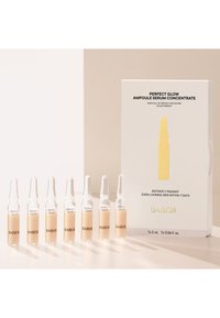 Ampoules en verre clair contenant un sérum jaune pâle, étiquetées "Perfect Glow", disposées à côté d'une boîte blanche avec du texte doré et des accents jaunes.