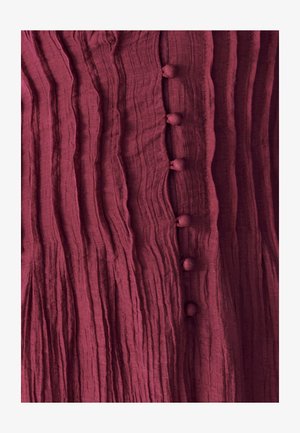 Bordová textúrovaná tkanina s vertikálnymi prehybmi a šiestimi okrúhlymi látkovými gombíkmi uprostred. Jemný lesk a mäkký vzhľad.